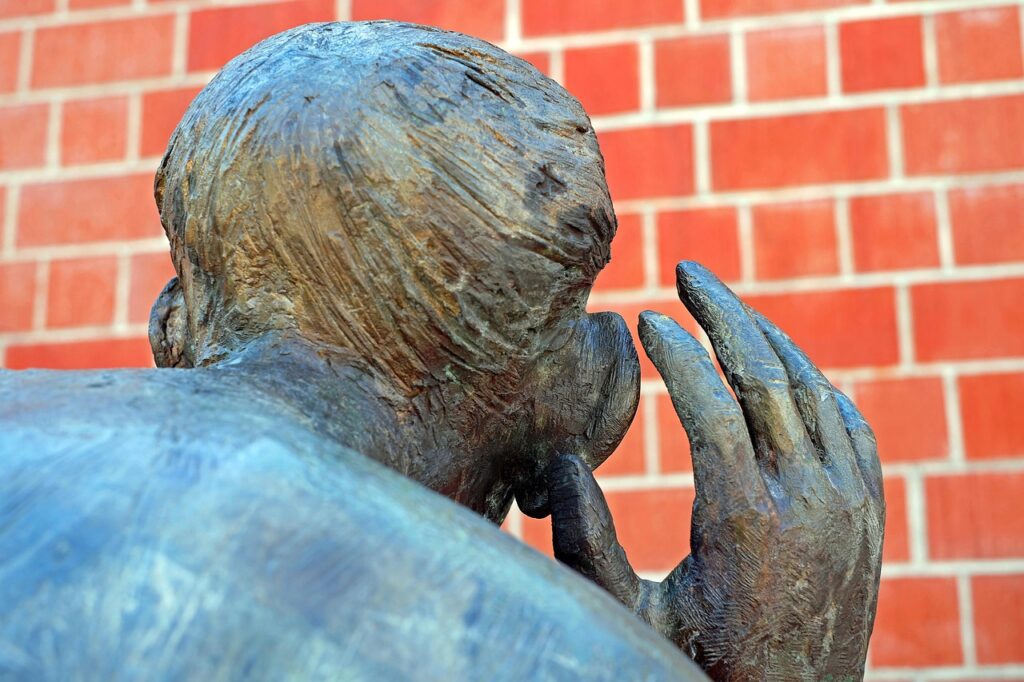 Sculpture d’un homme en bronze en train d’écouter, avec la main derrière l’oreille, devant un mur de briques rouges.