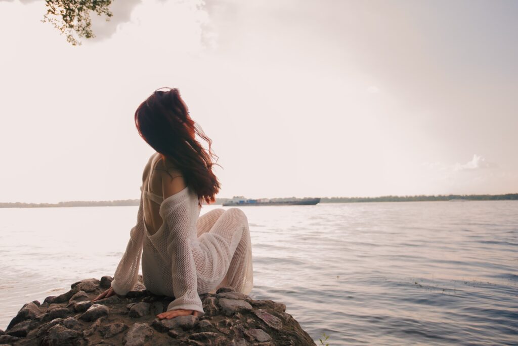 Femme assise sur un rocher au bord de l’eau, dans une posture contemplative et apaisée.